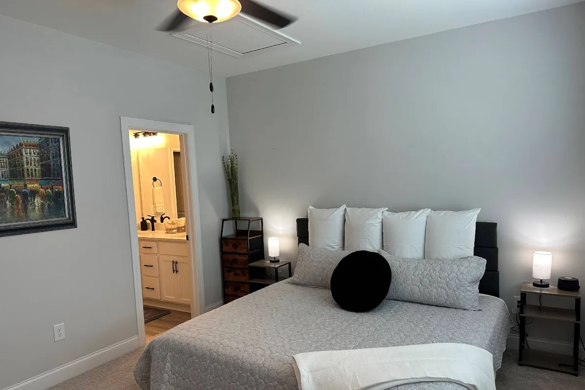 A bright, serene bedroom featuring a queen-sized bed adorned with decorative pillows, a piece of art above the headboard, and an inviting nightstand with lamps, creating a tranquil atmosphere ideal for unwinding.
