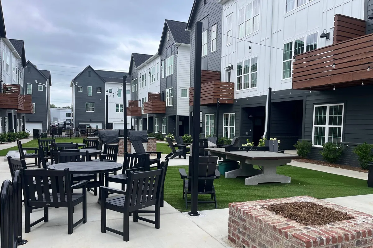 The image showcases the townhouse's modern exterior, featuring a stylish outdoor seating area set on a lush grassy lawn. The scene is inviting, with comfortable seating perfect for socializing or relaxing, and it is bathed in the warm glow of sunset. The backdrop hints at mountain views, blending city vibrance with natural beauty.