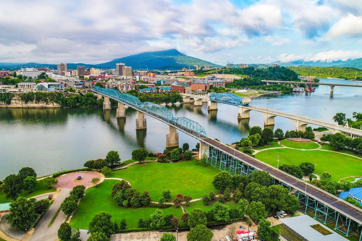 A broad view of Chattanooga’s downtown skyline, with the river winding through the city, bridges connecting neighborhoods, lush park areas, and the silhouette of mountains rising in the background. People can be seen walking, biking, and enjoying the vibrant urban landscape.