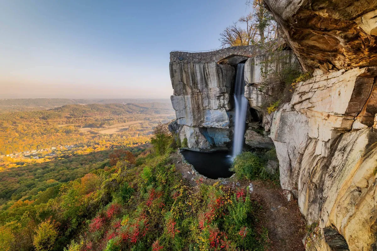 A stunning exterior shot capturing a tranquil waterfall cascading over natural rock formations with a broad valley view. Lush foliage surrounds the scene, creating a serene, inviting atmosphere that encourages outdoor exploration and relaxation after a productive workday.
