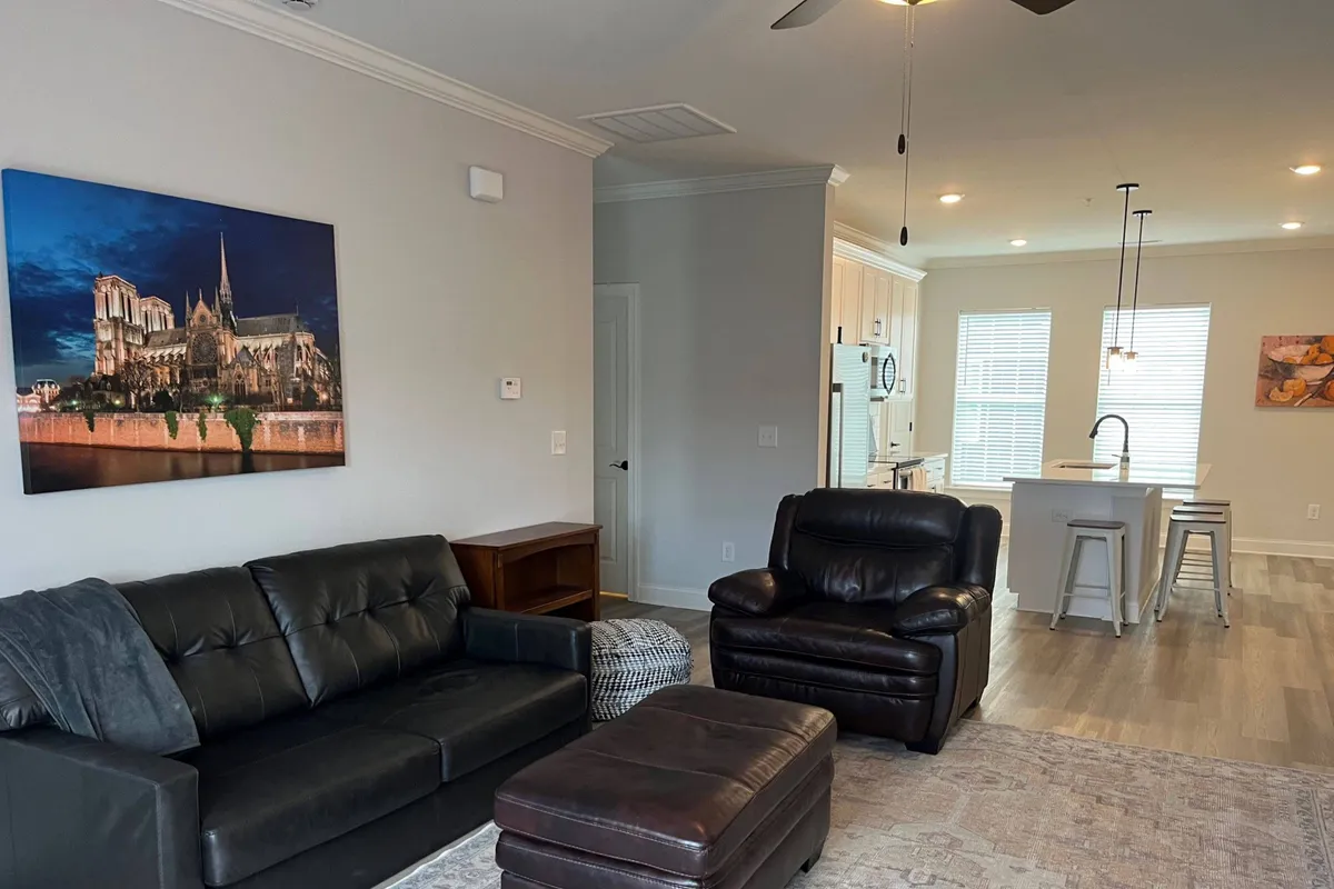 A modern, comfortable living room featuring a black leather sofa, brown leather armchair, and large wall art. The kitchen area is visible in the background, reinforcing the open-concept layout ideal for gatherings and relaxation.