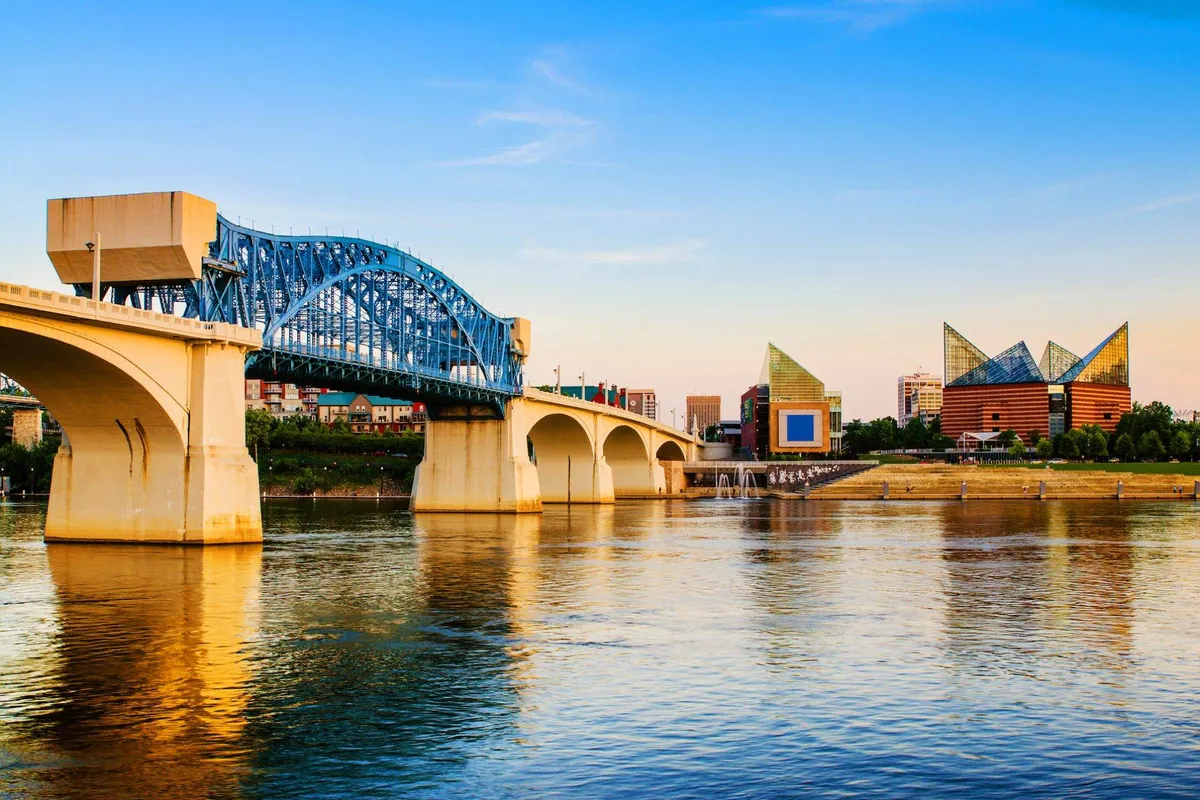 A tranquil view of a blue bridge spanning a river, with modern city buildings in the background and gentle sunset reflections on the water. The scene suggests easy connectivity to city attractions and scenic walks.