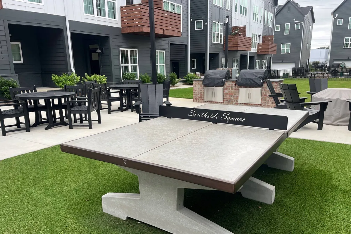 This image showcases an outdoor community space with a ping pong table, outdoor seating area, barbecue grills, and a green lawn. People can be seen engaging in lively activities such as playing ping pong, dining, and socializing.